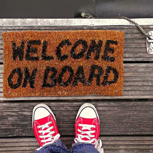 Das Bild zeigt ein paar rote Schuhe, die vor einer Fußmatte stehen, auf der geschrieben steht: Welcome on Board