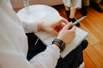 Im Vordergrund sind sie Hände einer Frau zu sehen, sie hat einen Stift in der Hand und ein Notizbuch liegt aufgeschlagen vor ihr. Im Vordergrund sind sie Hände einer Frau zu sehen, sie hat einen Stift in der Hand und ein Notizbuch liegt aufgeschlagen vor ihr.