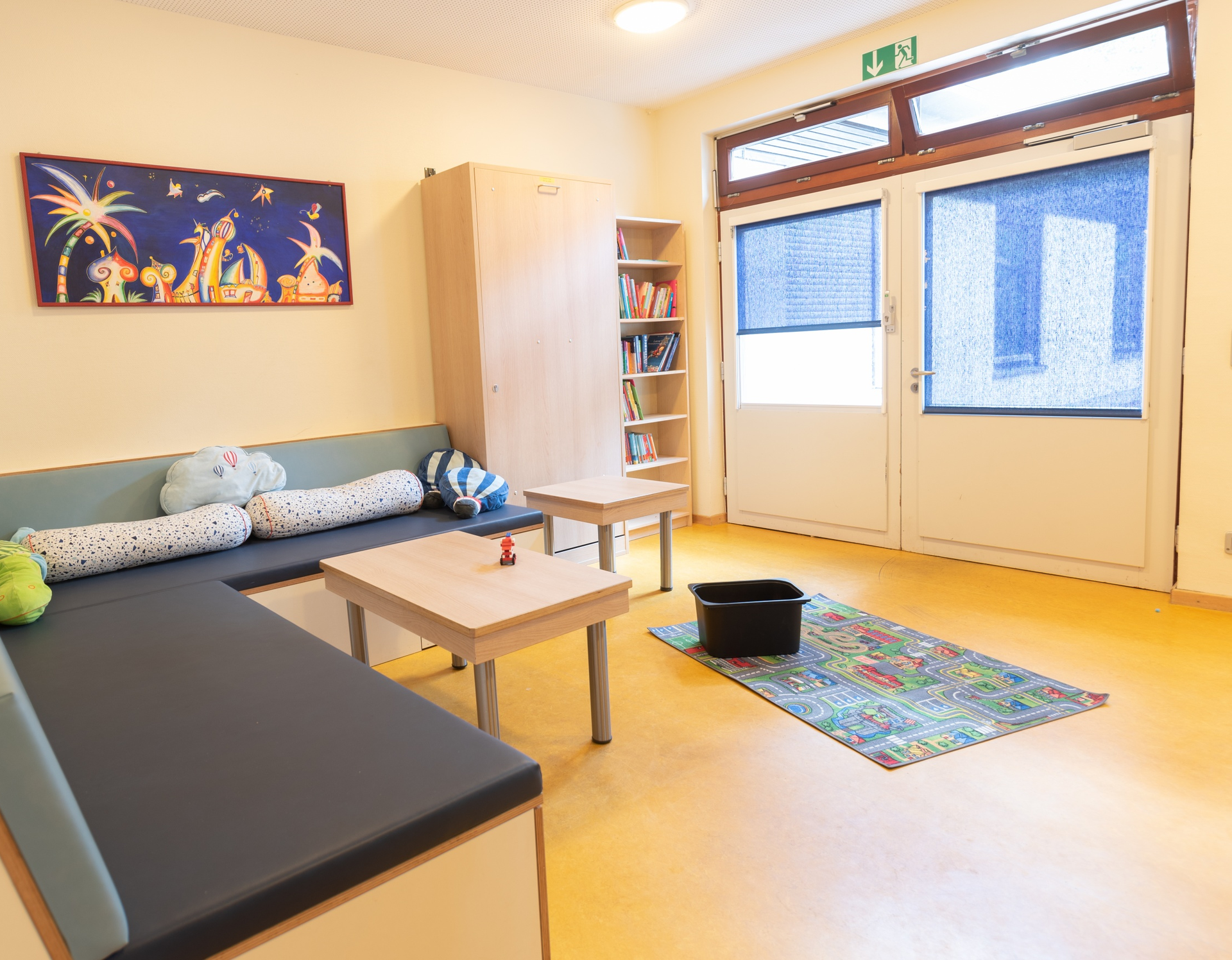Das Bild zeigt einen Wohnraum mit einem Sofa, zwei kleinen Tischen un einem Bücherragl. Auf dem Boden liegt ein Spielteppich.